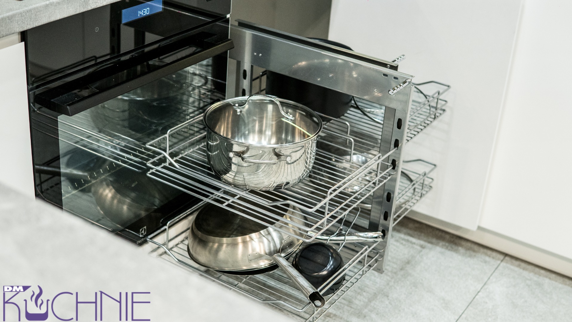 Wysuwana, metalowa szafka cargo w zabudowie kuchennej z garnkami i patelnią, widok na piekarnik z wyświetlaczem czasu.