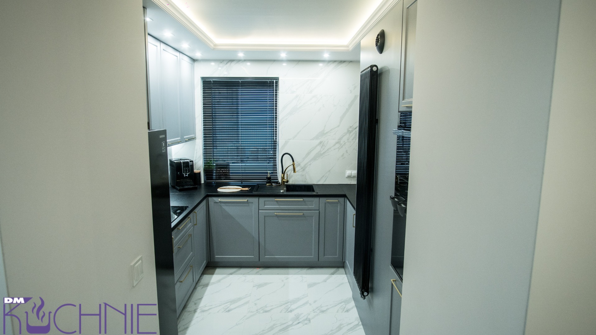 Nowoczesna kuchnia w odcieniach szarości z czarnym blatem i złotą armaturą, marmurowe płytki na ścianie i podłodze, widok zza ściany.