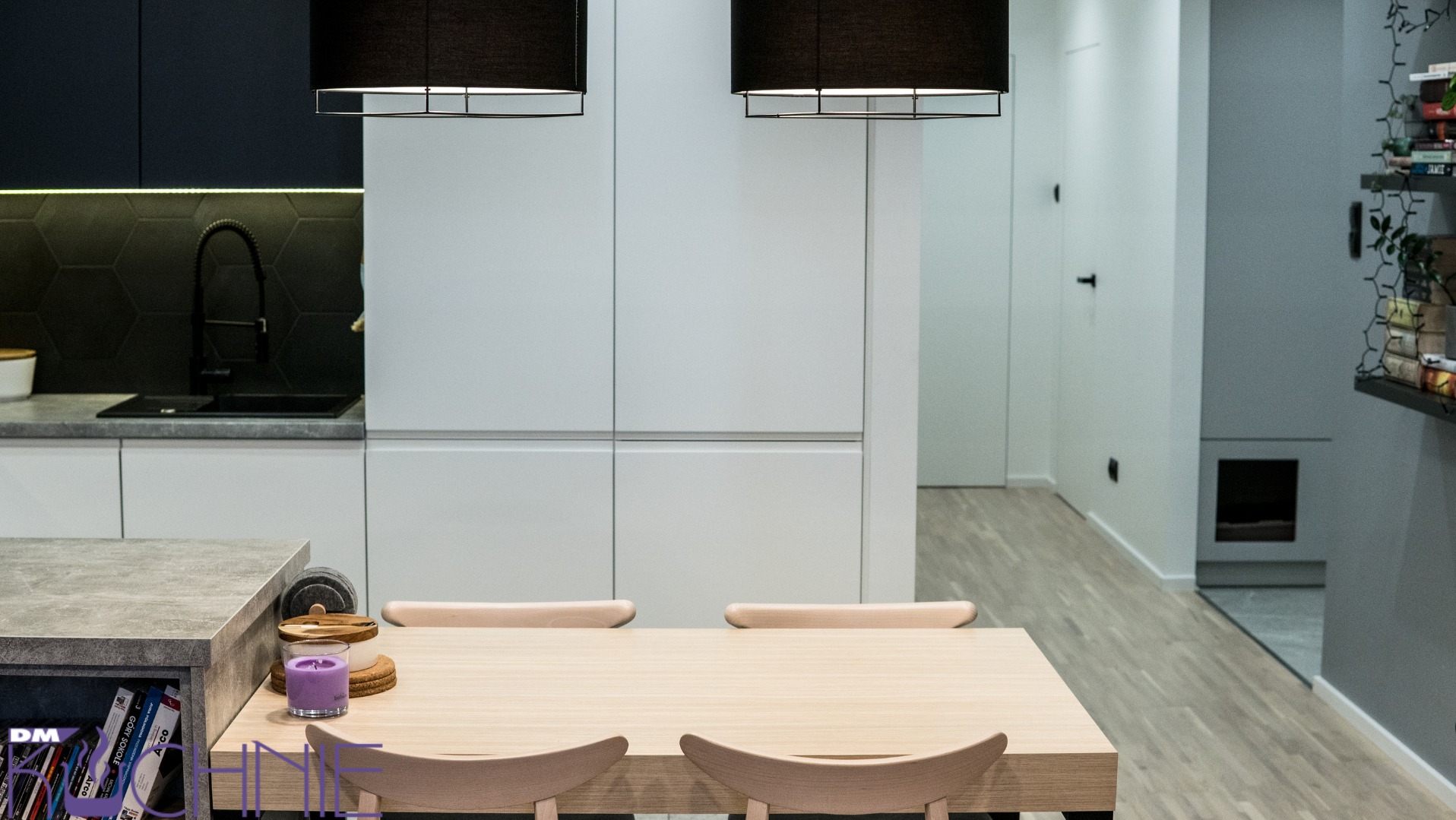 Nowoczesna kuchnia z białymi szafkami, czarną armaturą i heksagonalnymi płytkami, oświetlona dwoma wiszącymi lampami nad drewnianym stołem z krzesłami, uzupełniona fioletową świecą i książkami.