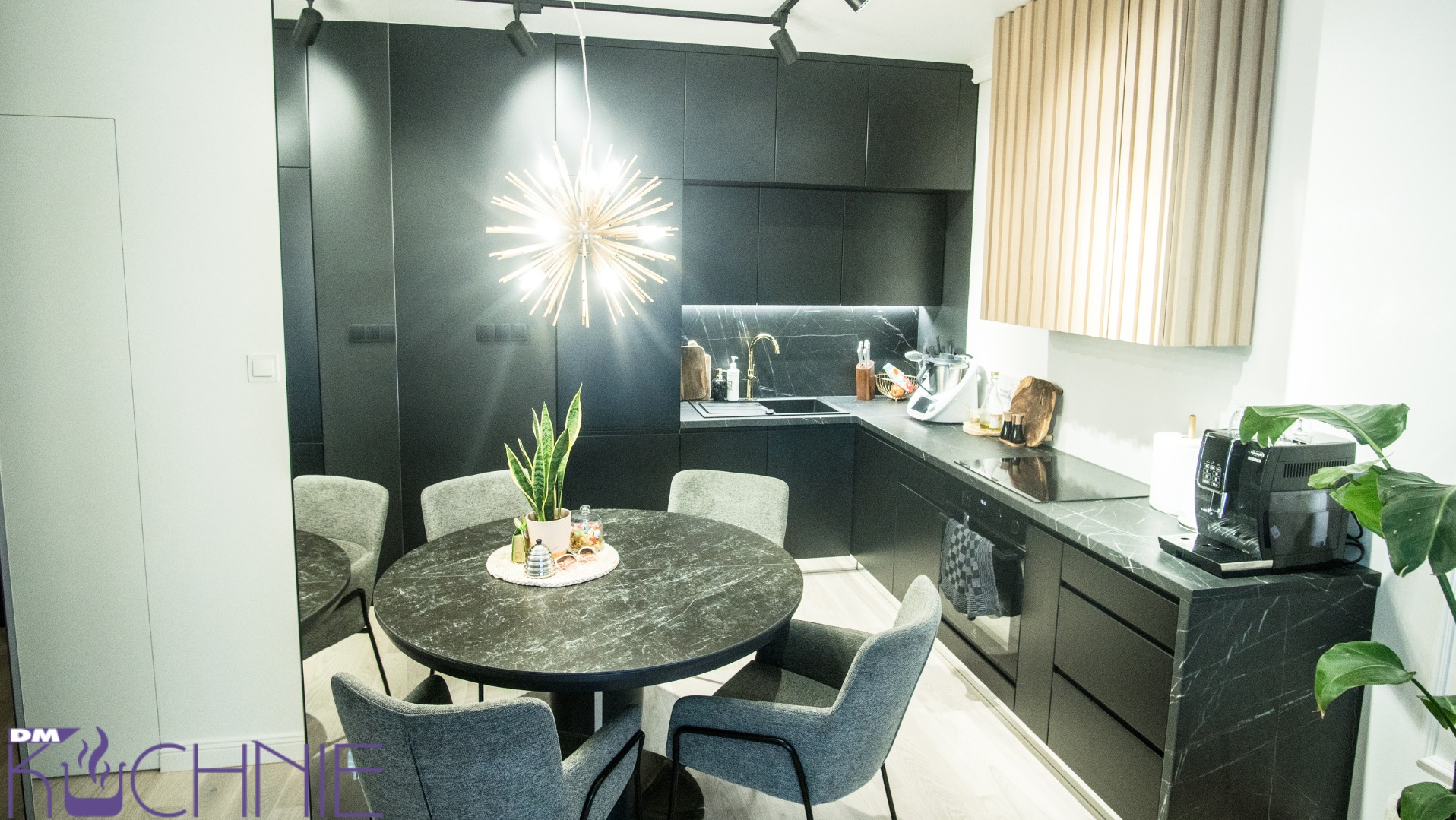 Nowoczesna, czarna kuchnia z okrągłym stołem z blatem imitującym marmur i szarymi krzesłami. Nad stołem wisi designerska lampa. W tle widoczny fragment zabudowy kuchennej z czarnymi szafkami...