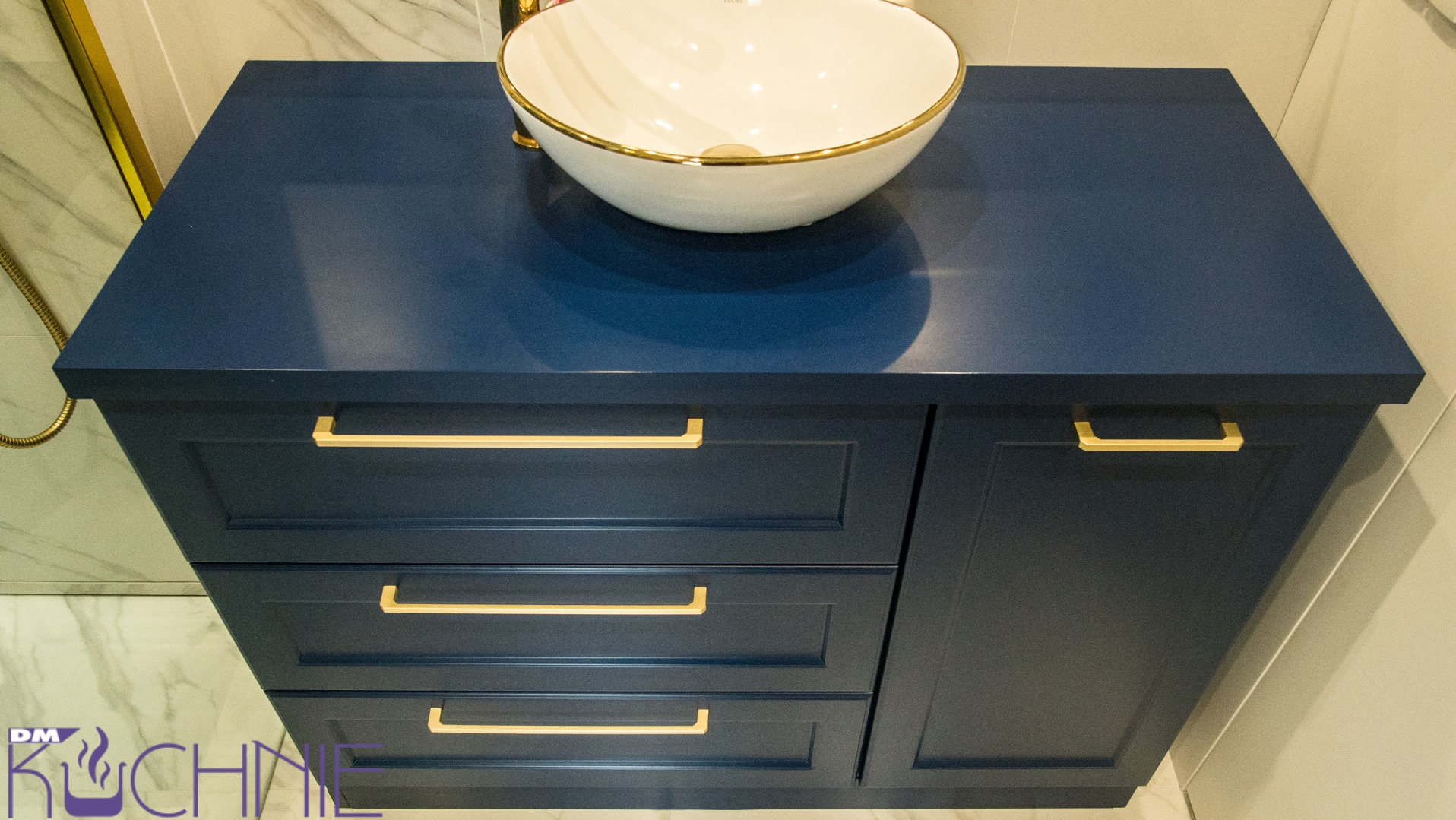 Niebieska szafka łazienkowa z trzema szufladami i umywalką nablatową z złoconym rantem, widoczne złote uchwyty i fragment marmurowej ściany