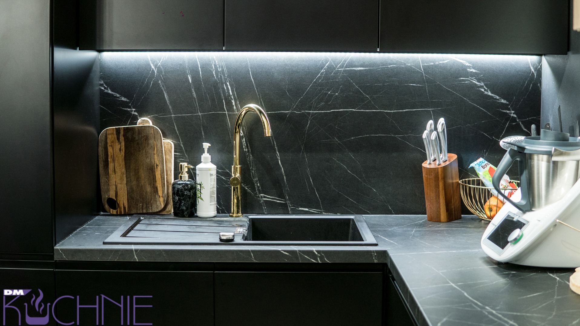 Czarne, matowe szafki kuchenne z blatem imitującym marmur, złota bateria, deski do krojenia, dozowniki mydła, blok z nożami i robot kuchenny Thermomix w nowoczesnej kuchni.