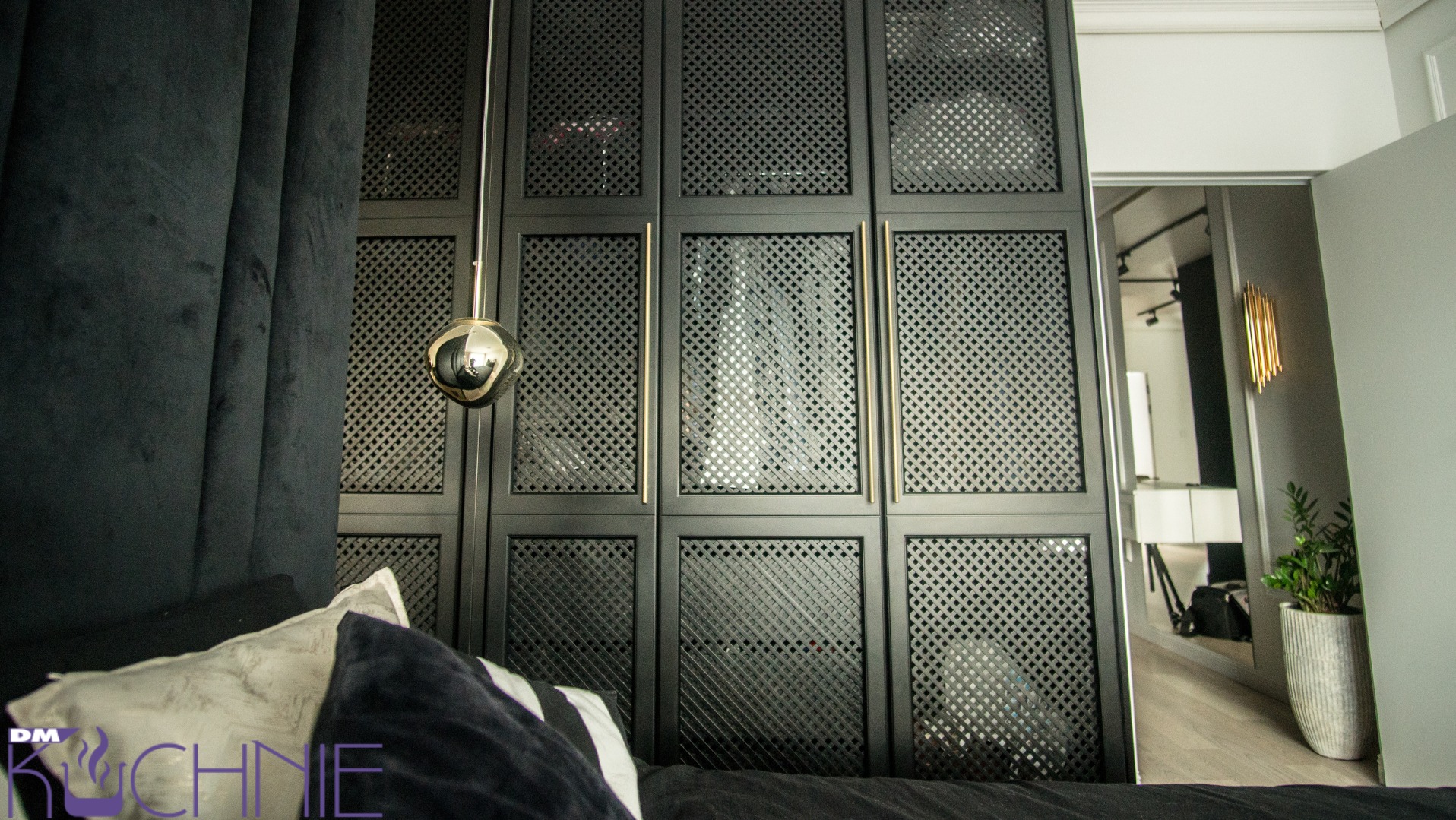 Czarna szafa wnękowa z perforowanymi drzwiami i złotymi uchwytami, widoczna z perspektywy łóżka, z lampą wiszącą w kształcie kuli i fragmentem zasłony.