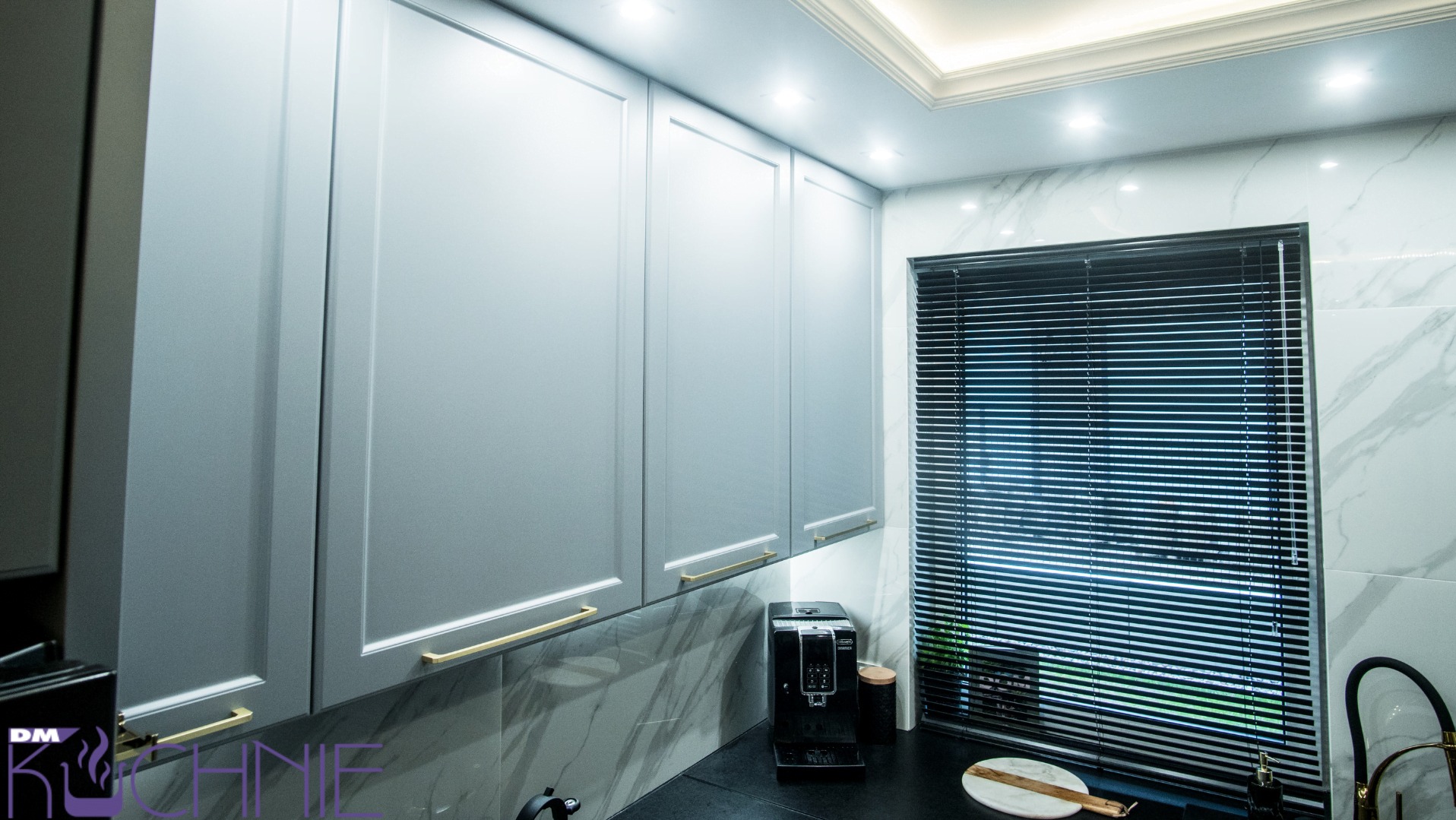 Szare szafki kuchenne z frezowanymi frontami i złotymi uchwytami, marmurowe płytki na ścianie, ekspres do kawy na blacie, widok z dołu na oświetlenie sufitowe.