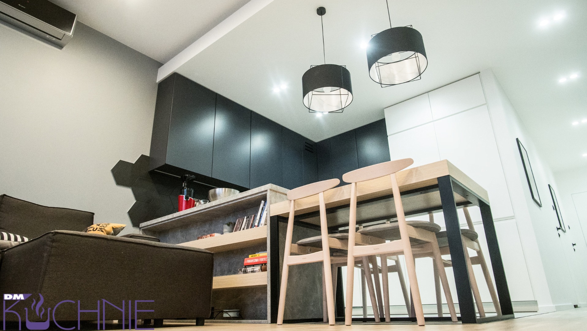 Częściowy widok nowoczesnej kuchni z czarnymi szafkami, drewnianym stołem i krzesłami, oświetleniem sufitowym i dekoracyjną ścianą z heksagonów, widzianej z niskiego kąta