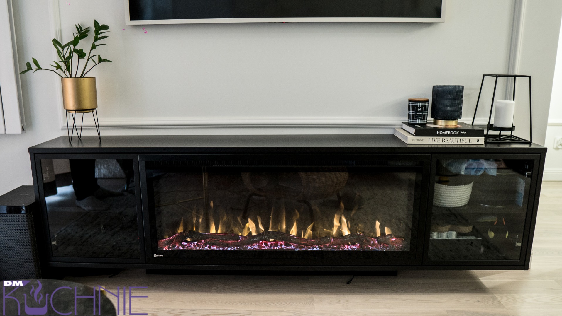 Czarna szafka RTV z elektrycznym kominkiem i szklanymi drzwiczkami, dekoracje na blacie, roślina w złotej doniczce, widoczne odbicie osoby w szybie.