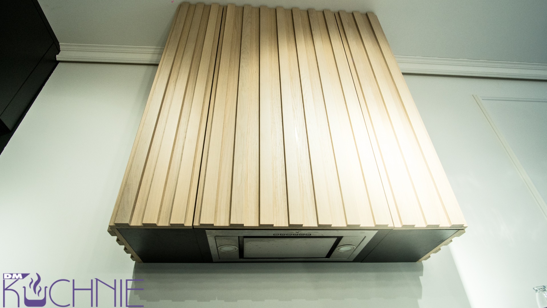 Nowoczesny okap kuchenny z obudową z drewnianych listewek w jasnym odcieniu, zintegrowany z czarną szafką, widoczny na tle białej ściany z dekoracyjną listwą przysufitową.