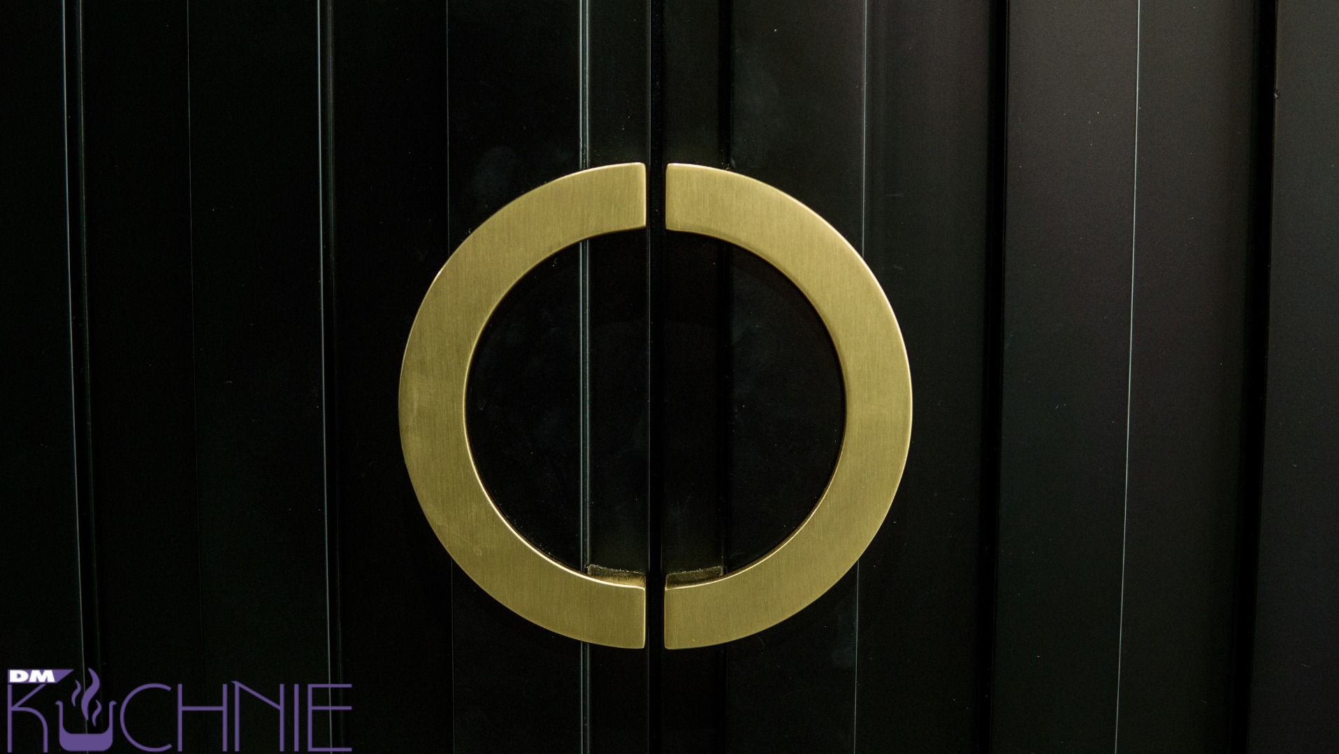 Czarne fronty szafek z frezowanymi pionowymi liniami i okrągłym, złotym uchwytem dwuczęściowym.