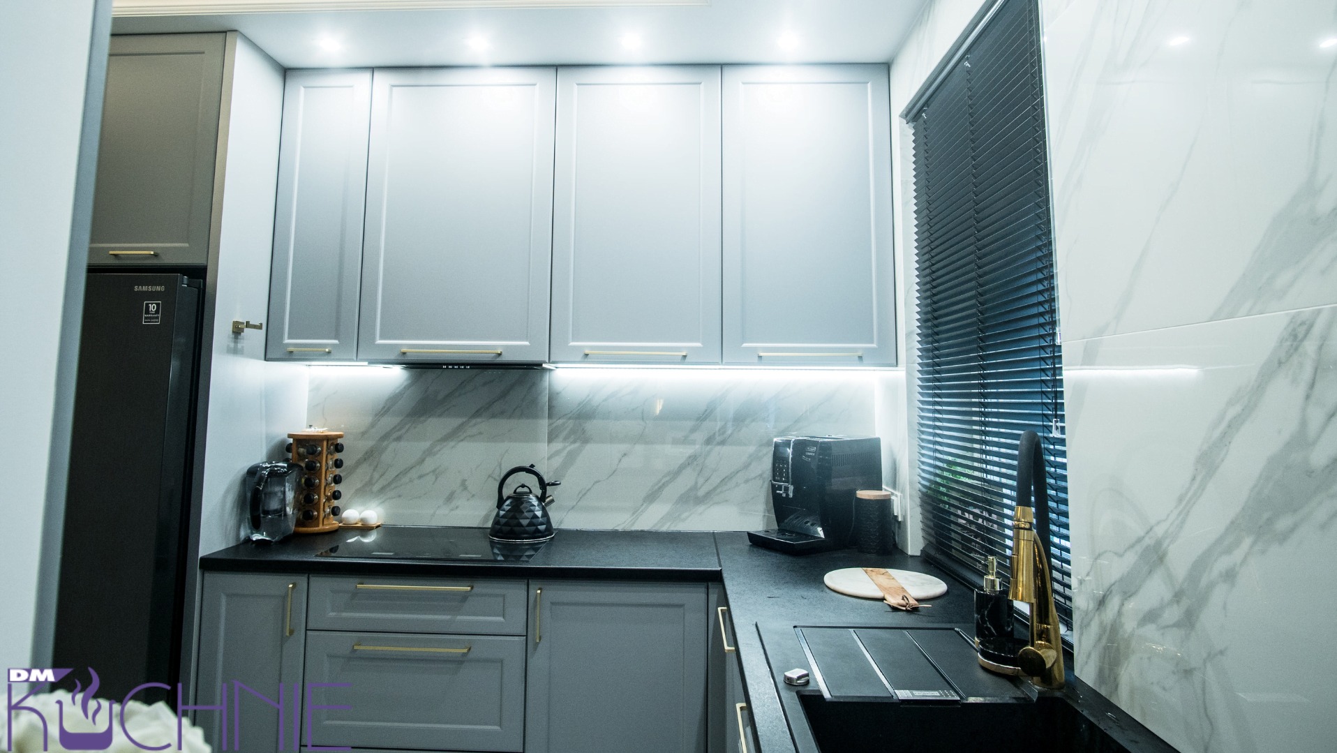 Szara kuchnia z marmurowym backsplash, złotymi uchwytami i czarnym zlewem pod oknem z żaluzjami.