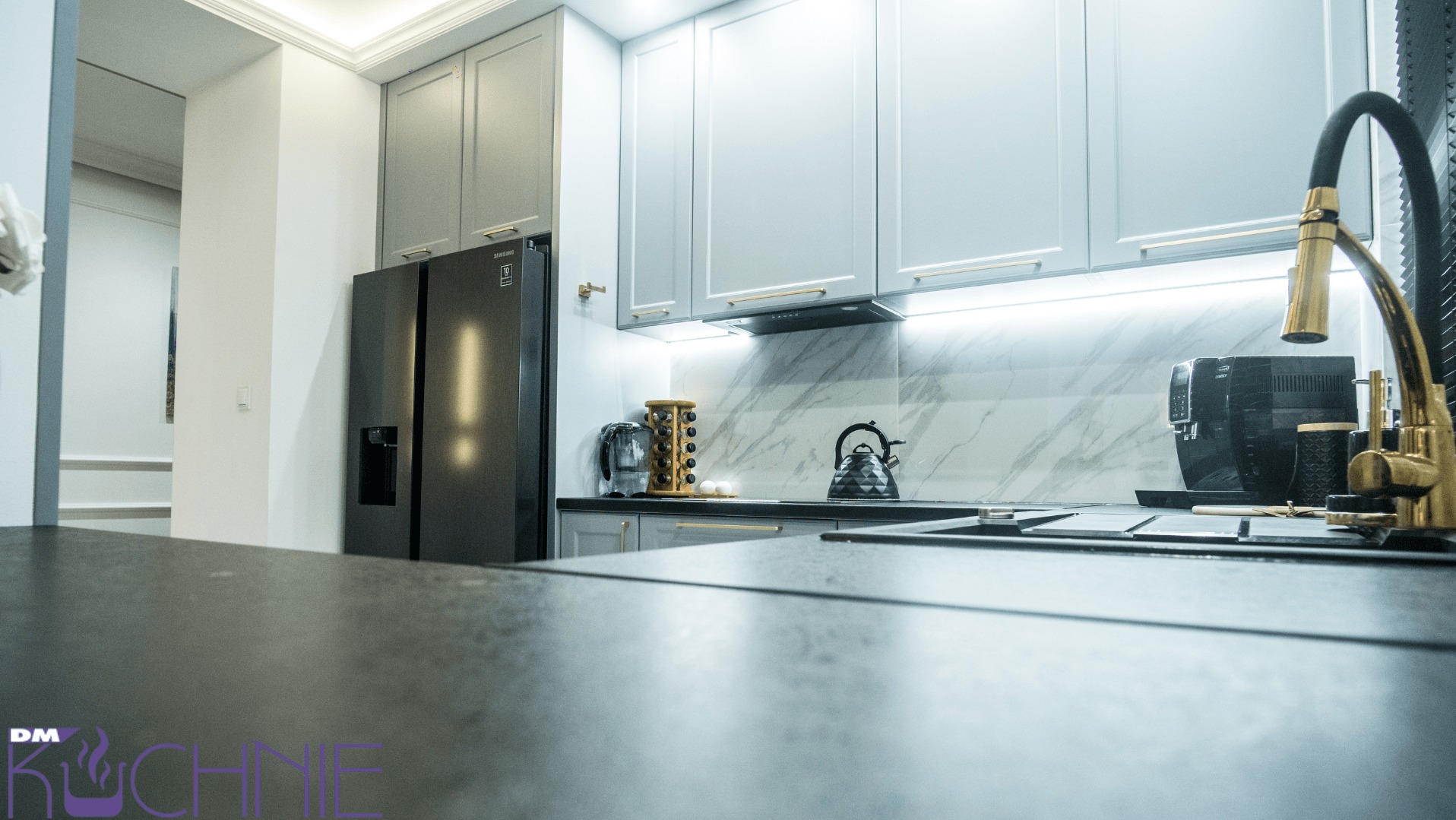 Nowoczesna kuchnia z szarymi szafkami, marmurowym backsplashiem, czarną lodówką Samsung i złotą baterią, widziana z perspektywy blatu. W tle ekspres do kawy i czajnik.