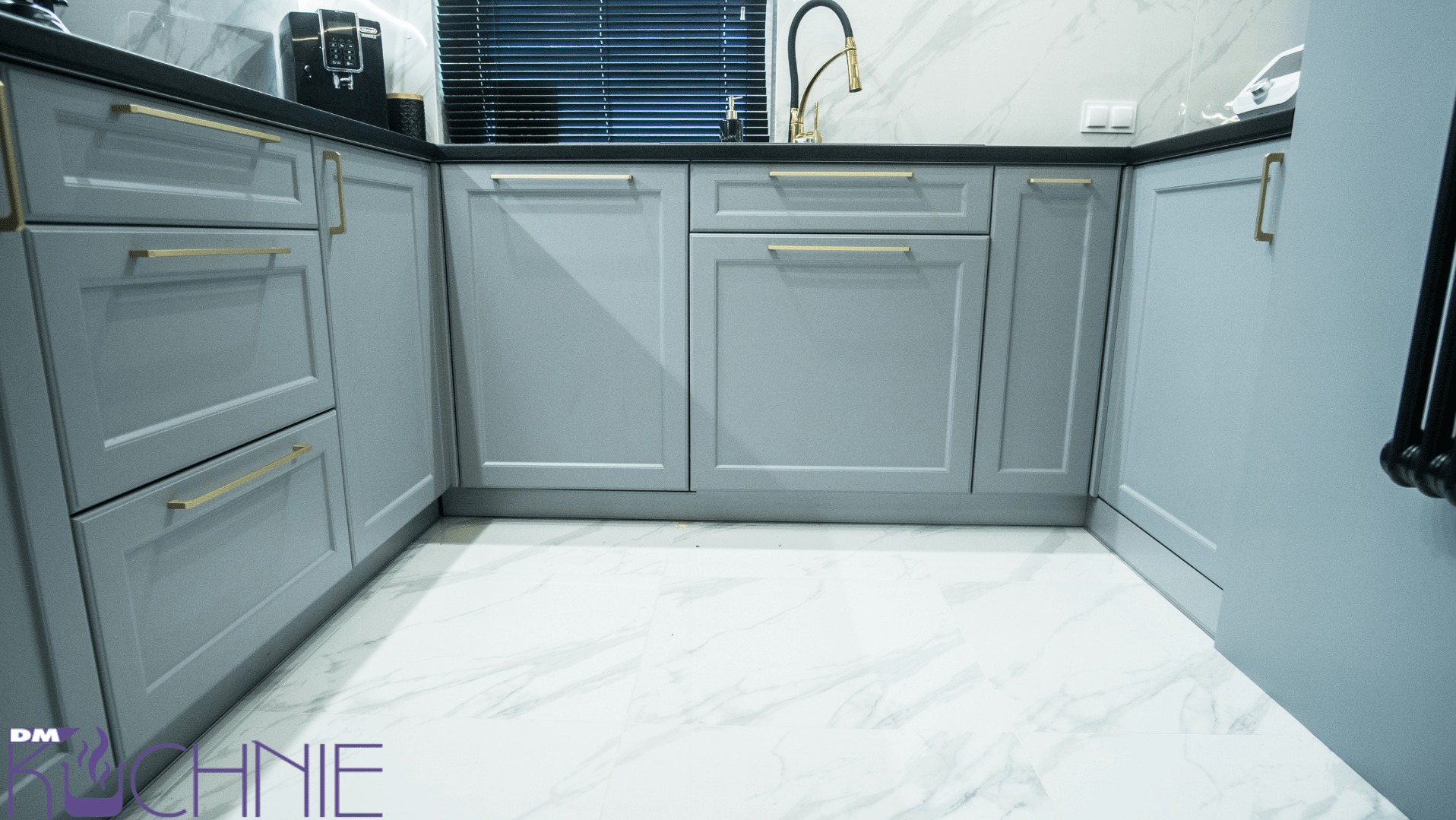 Szare szafki kuchenne z złotymi uchwytami na tle marmurowej ściany i podłogi, widok z dołu.