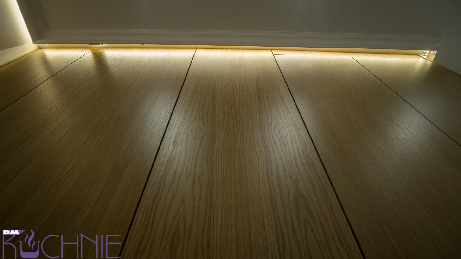 Fronty szafek kuchennych z naturalnego drewna, oświetlenie LED podszafkowe, widok od dołu, logo w lewym dolnym rogu.