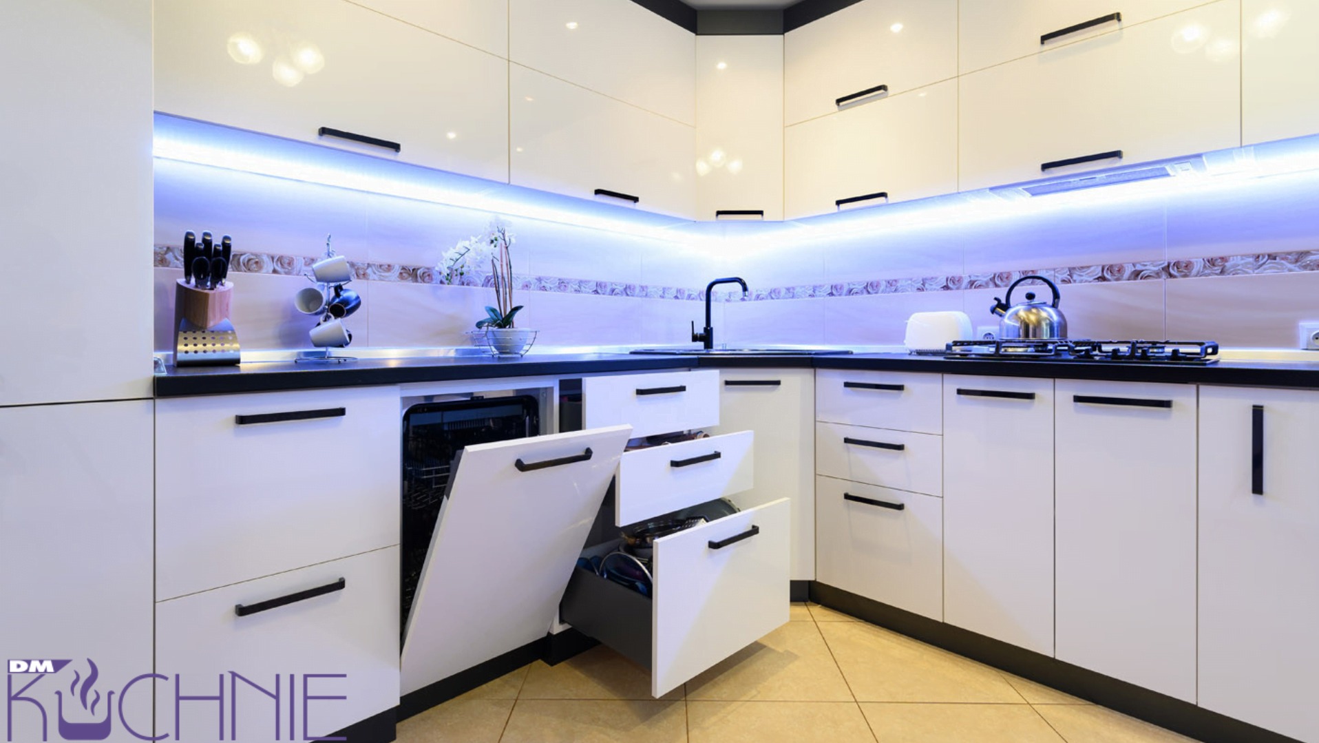 Nowoczesna, biała kuchnia z czarnymi akcentami, oświetleniem LED pod szafkami, otwartą zmywarką i wysuwanymi szufladami, widoczny fragment płyty gazowej i czajnika.