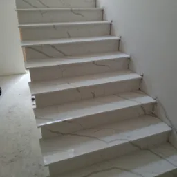 Białe schody z efektem marmuru, widoczne z dołu, z delikatnym oświetleniem z okna na górze. Wykończenie w trakcie montażu, widoczne zabezpieczenia na krawędziach.