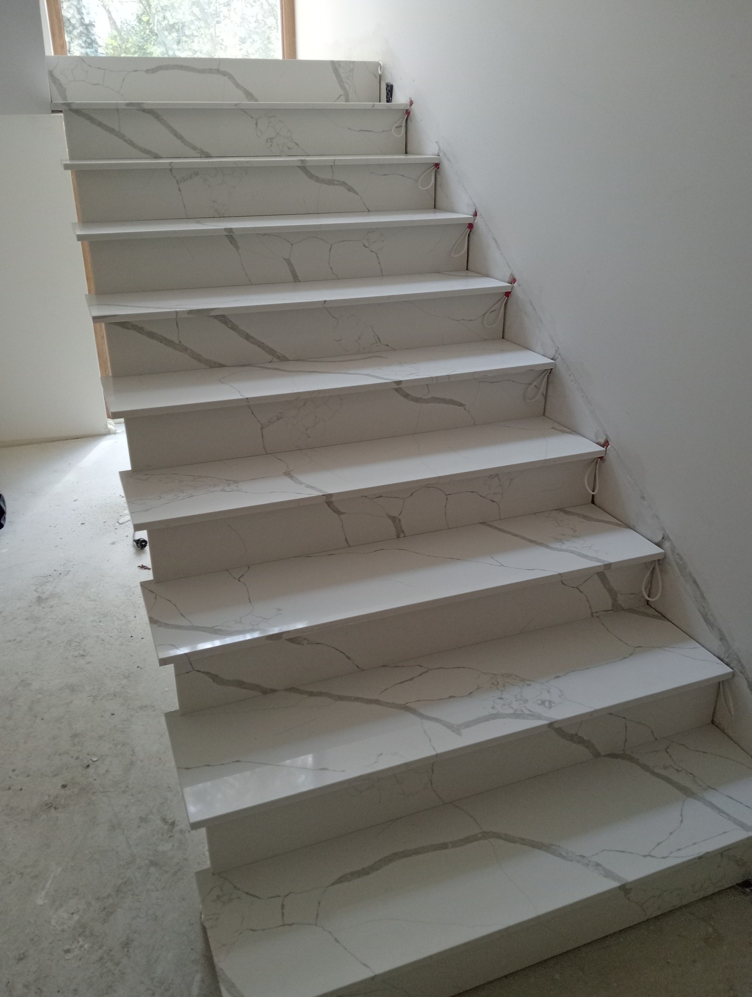 Białe schody z efektem marmuru, widoczne z dołu, z delikatnym oświetleniem z okna na górze. Wykończenie w trakcie montażu, widoczne zabezpieczenia na krawędziach.