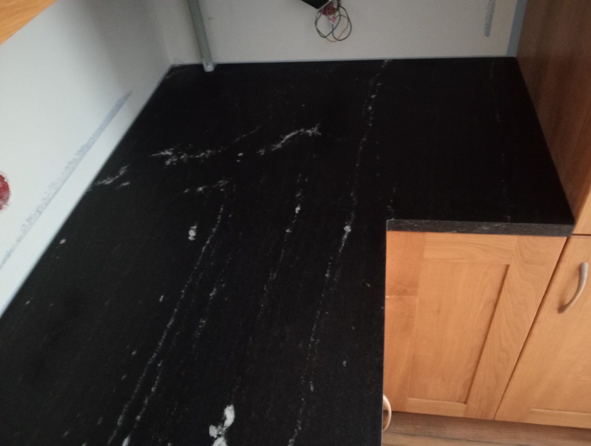 Czarny blat kamienny z subtelnym, białym użyleniem, zamontowany w kuchni obok drewnianych szafek. Widoczne fragmenty ściany i instalacji.