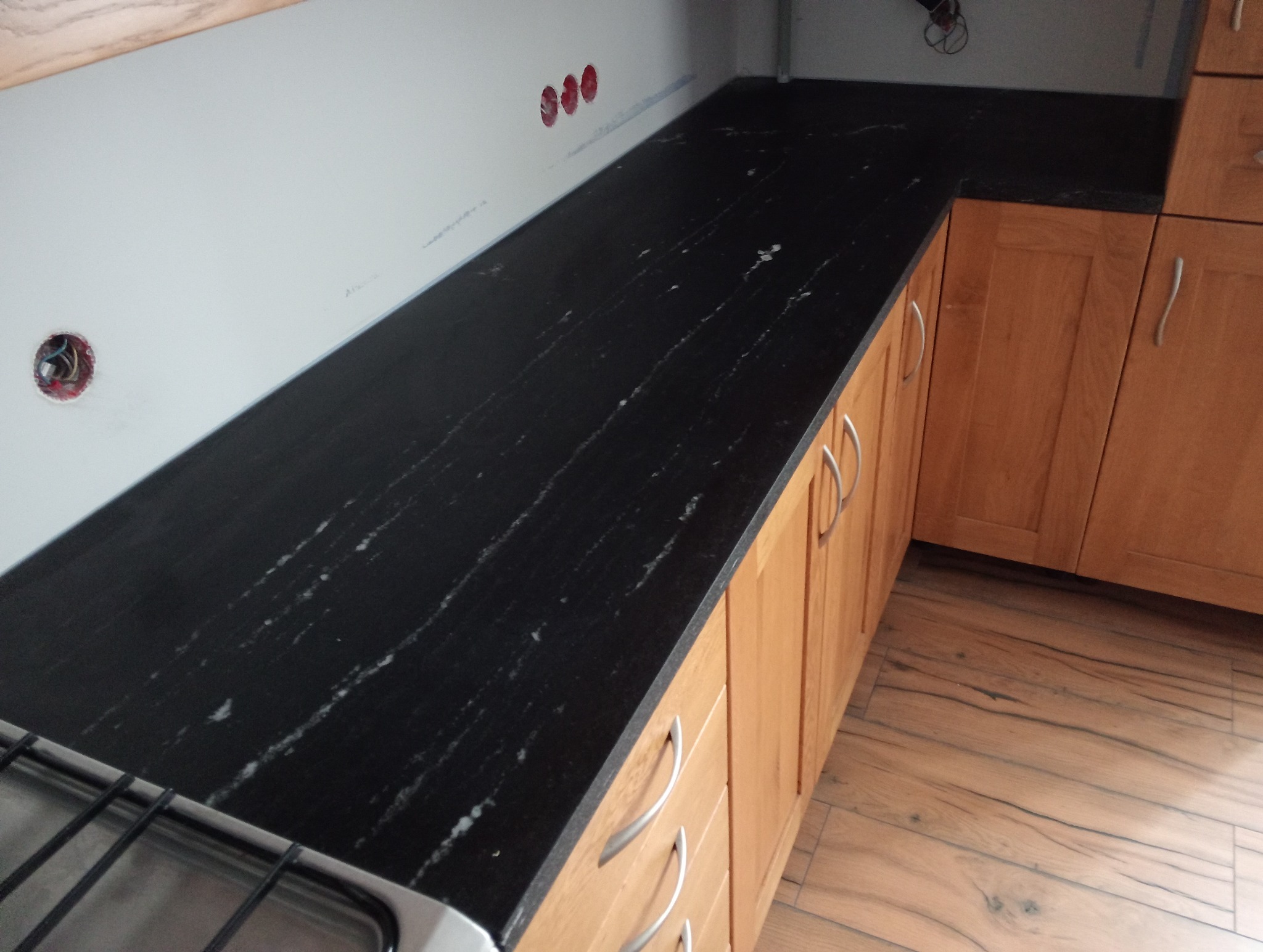 Czarny blat kamienny z jasnymi żyłkami, zamontowany w kuchni z drewnianymi szafkami. Widoczne gniazdka elektryczne na ścianie.