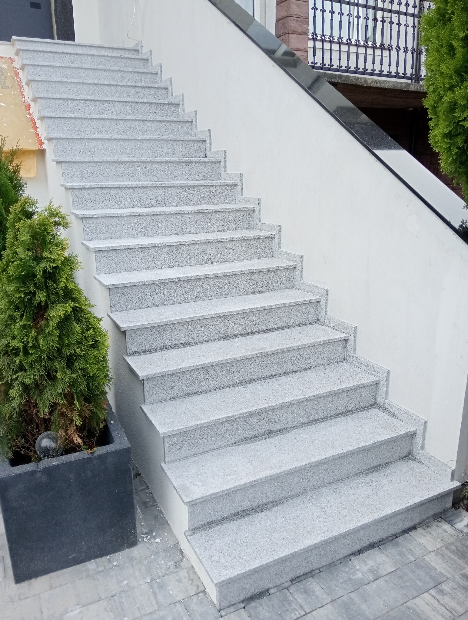 Zewnętrzne schody z jasnego granitu prowadzące do domu, z donicą z rośliną iglastą po lewej stronie. Widoczny fragment elewacji i balustrady.