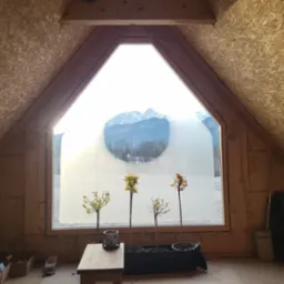 Poddasze z oknem w kształcie domu, przez kt&oacute;re widać ośnieżone g&oacute;ry i zaśnieżone pole, a na parapecie stoją cztery małe drzewka w doniczkach.