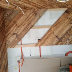 Wnętrze w trakcie wykańczania: drewniana boazeria na skośnym suficie i ścianach, widoczne przewody elektryczne w peszlach, fragment ściany z bloczków i płyta gipsowo-kartonowa oparta o nią.