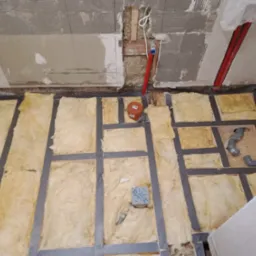 Przygotowanie podłoża pod podłogę: izolacja z wełny mineralnej między legarami, widoczne rury instalacji wodnej i kanalizacyjnej, surowy stan ścian.
