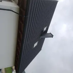 Ciemnoszary dach z blachy na nowoczesnym domu z oknami dachowymi i orynnowaniem, widziany z dołu pod pochmurnym niebem.