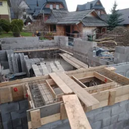 Budowa fundamentów domu jednorodzinnego z bloczków betonowych, widoczne szalunki i zbrojenie, w tle zabudowa w stylu góralskim.
