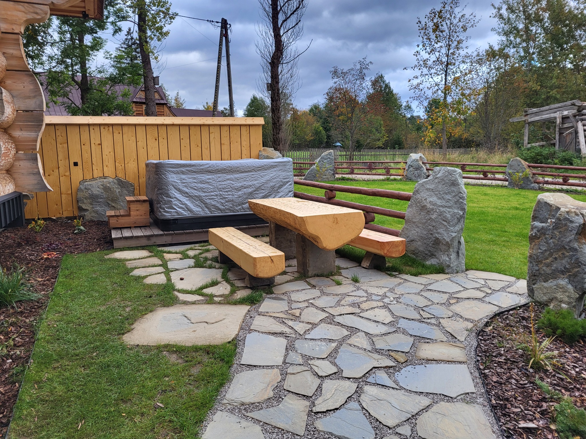 Zewnętrzne jacuzzi przykryte plandeką, obok drewniany stół i ławki z bali, kamienny chodnik, trawnik i ogrodzenie z bali na tle drzew.