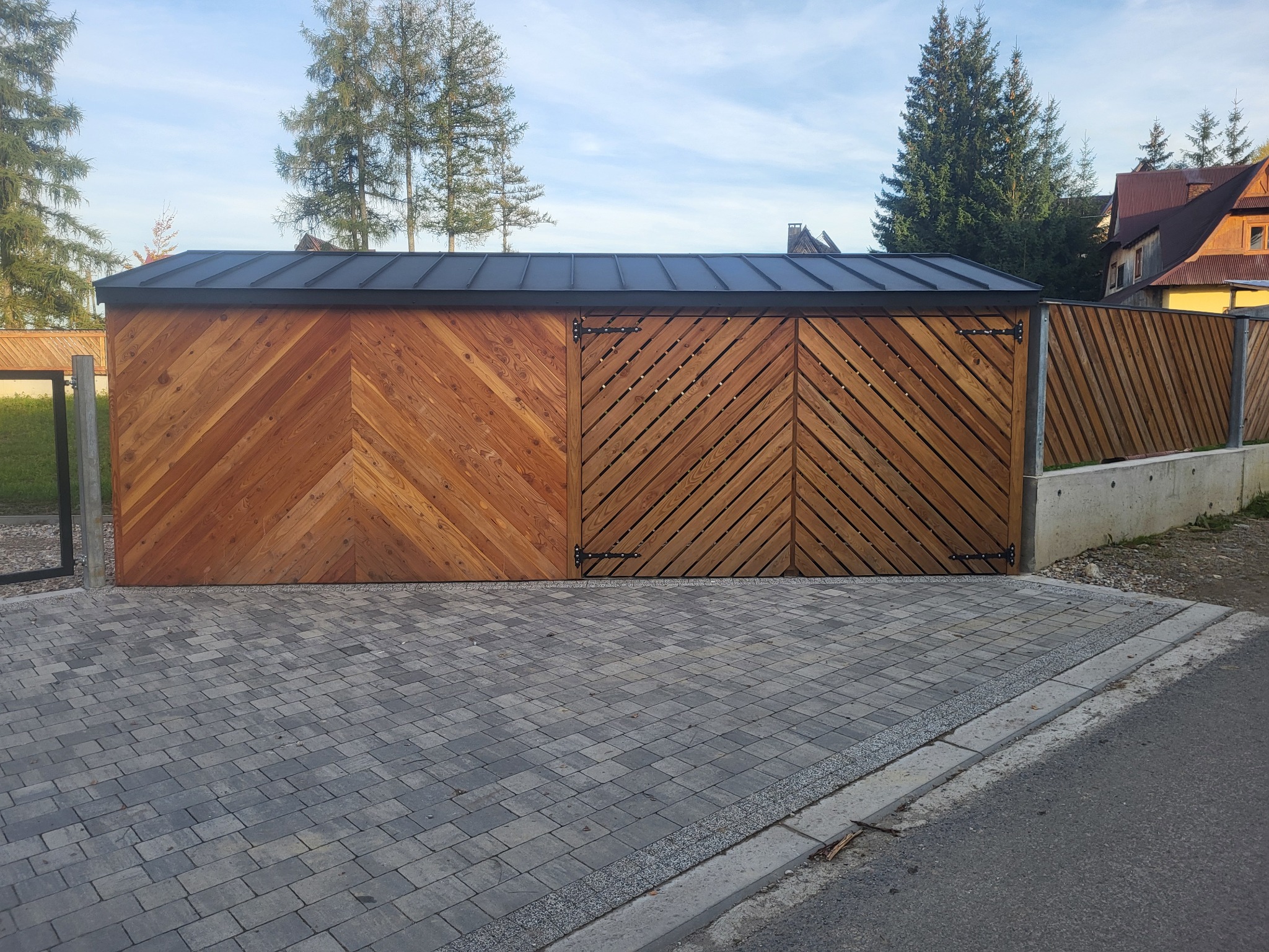 Drewniana konstrukcja z dwoma rodzajami ułożenia desek w jodełkę, z czarnym dachem, widoczna z zewnątrz na tle brukowanej drogi i zieleni.