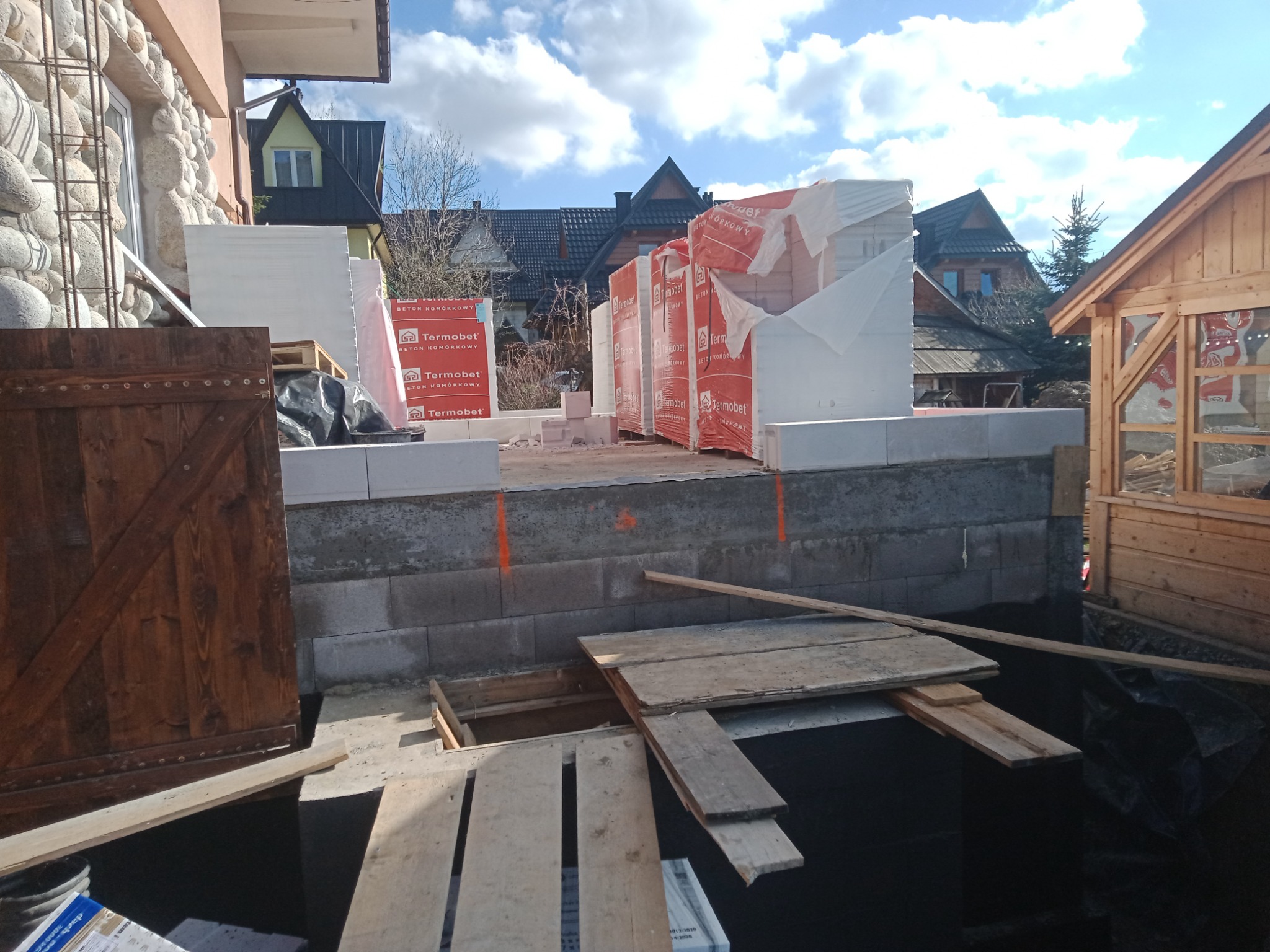 Budowa tarasu: widok na fundament z bloczków betonowych, przygotowane podłoże z czarną folią, palety z bloczkami Termobet w tle, drewniana konstrukcja altany po prawej, dom z kamienną elewacją po...