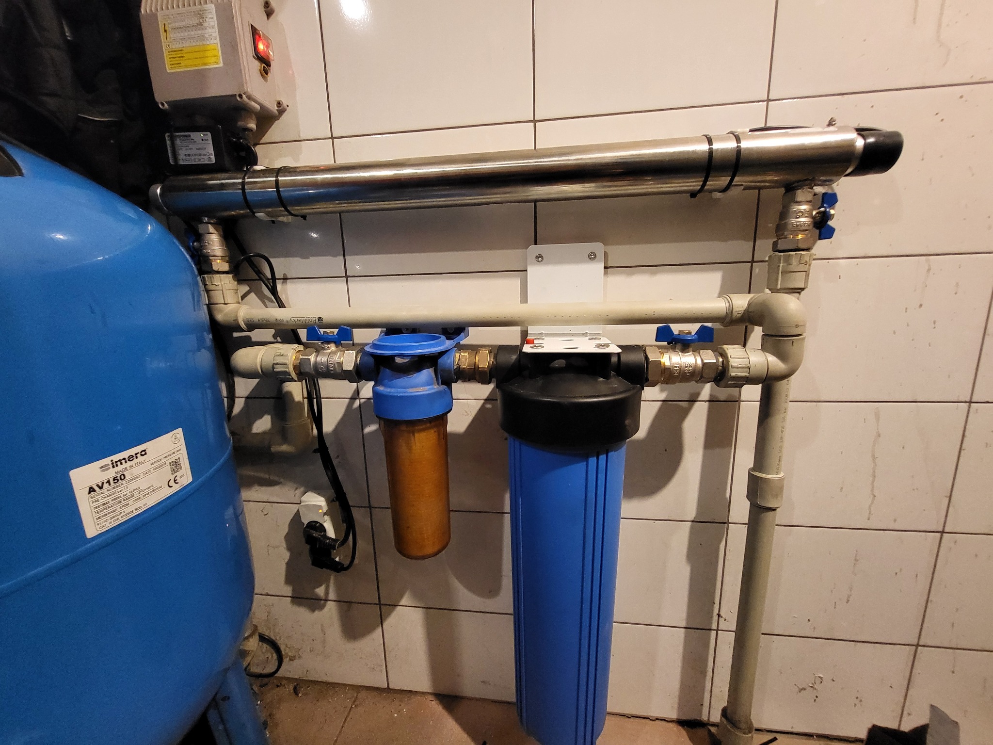 Instalacja uzdatniania wody z niebieskim zbiornikiem ciśnieniowym, filtrami mechanicznymi i lampą UV, zamontowana na tle białych, kwadratowych płytek ceramicznych.