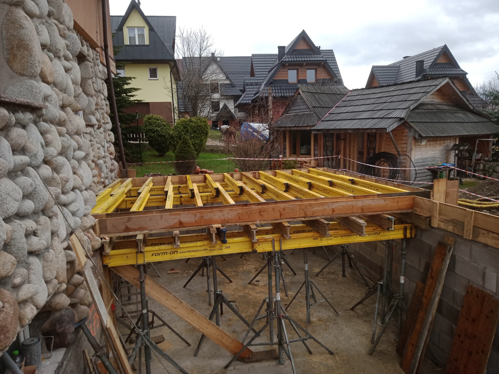 Żółte podpory i drewniane szalunki przygotowane pod wylewkę stropu w nowo budowanym domu, widok na kamienną ścianę i okoliczną zabudowę w tle.