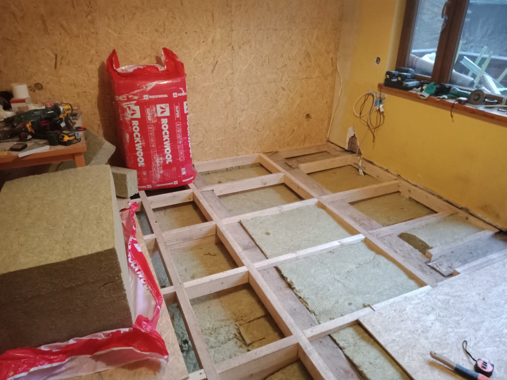 Ocieplanie podłogi: drewniana konstrukcja legarów z wełną mineralną Rockwool, widoczna paczka wełny, ściany wykończone płytą OSB, narzędzia na parapecie okna.