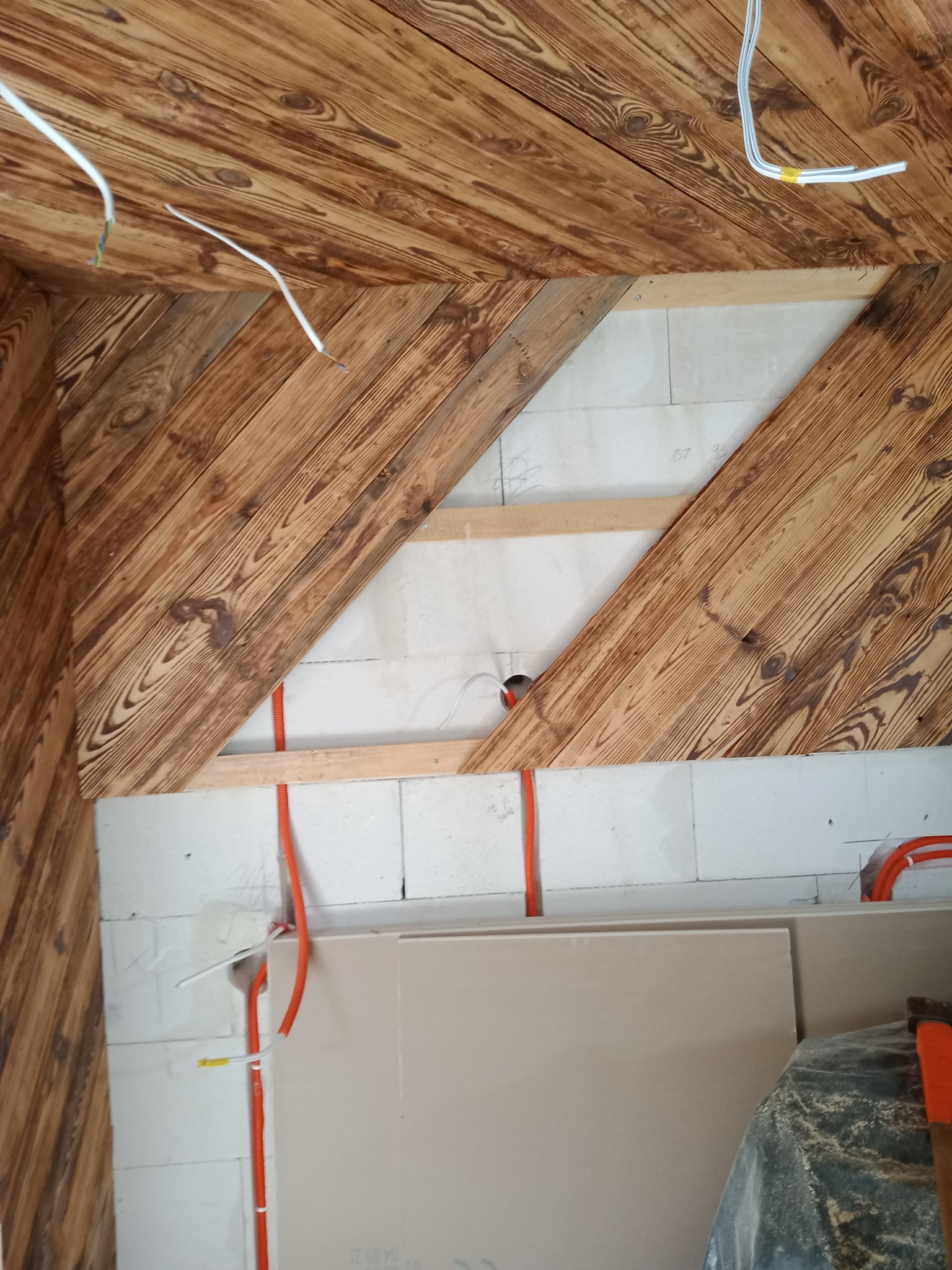 Wnętrze w trakcie wykańczania: drewniana boazeria na skośnym suficie i ścianach, widoczne przewody elektryczne w peszlach, fragment ściany z bloczków i płyta gipsowo-kartonowa oparta o nią.