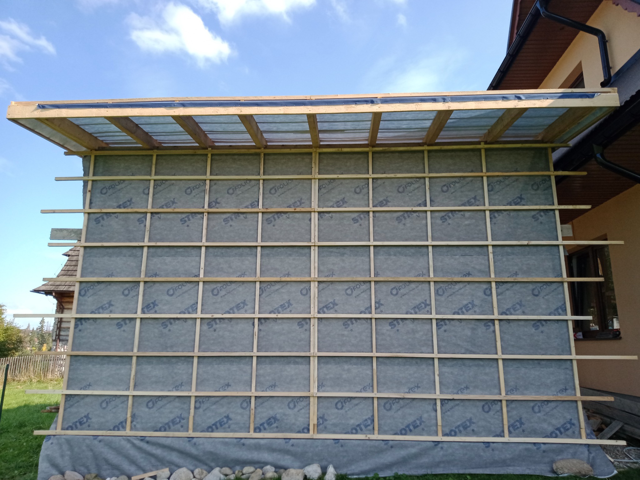 Konstrukcja drewniana pod elewację z folią wiatroizolacyjną, widoczne elementy mocujące, fragment dachu i ściany budynku w tle.
