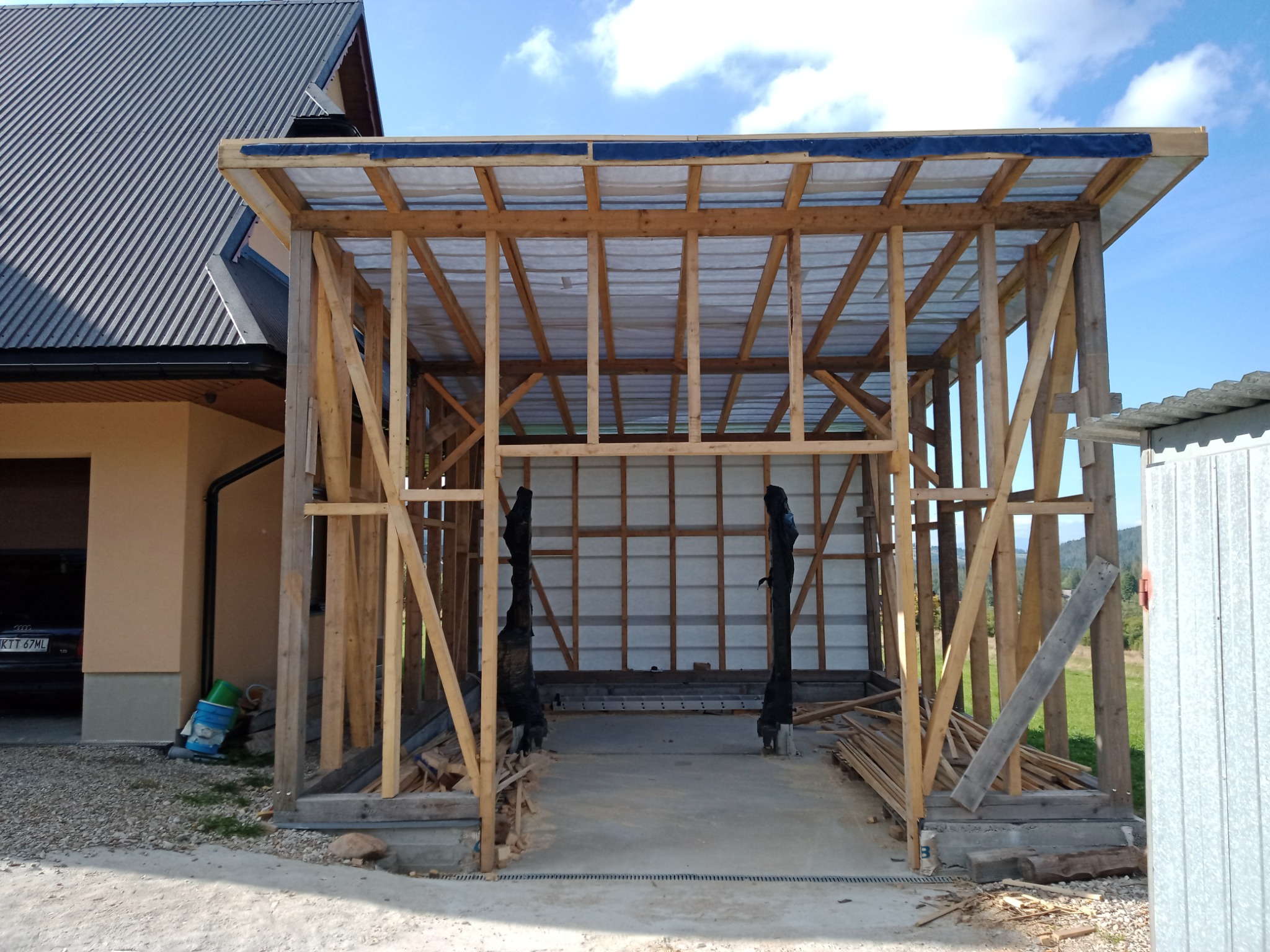 Drewniana konstrukcja zadaszenia w trakcie budowy, widoczne elementy szkieletu i tymczasowe zabezpieczenie dachu, w tle budynek mieszkalny i błękitne niebo.