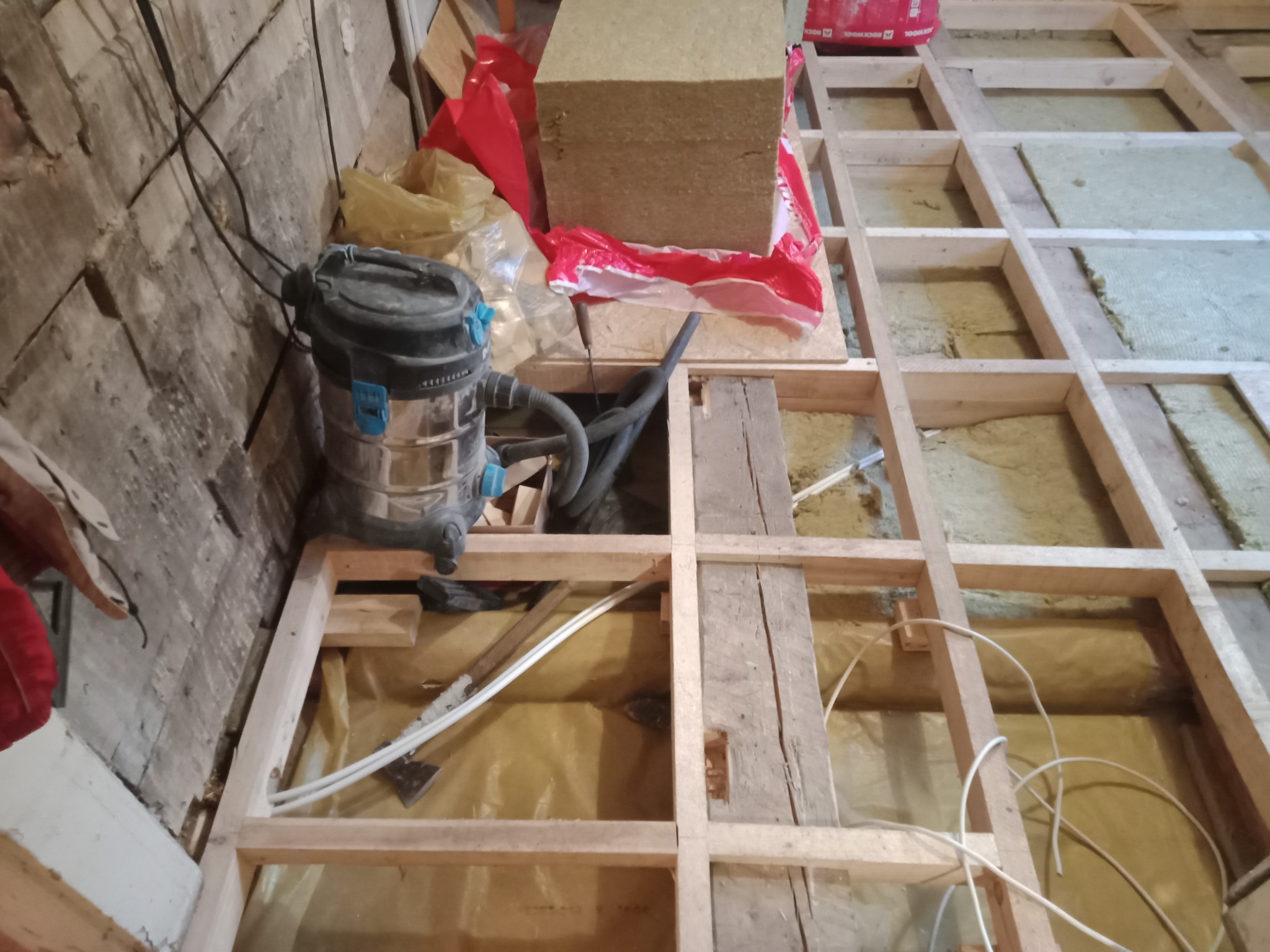 Drewniany szkielet podłogi z izolacją, widoczny odkurzacz przemysłowy i materiały izolacyjne w czerwonym worku.