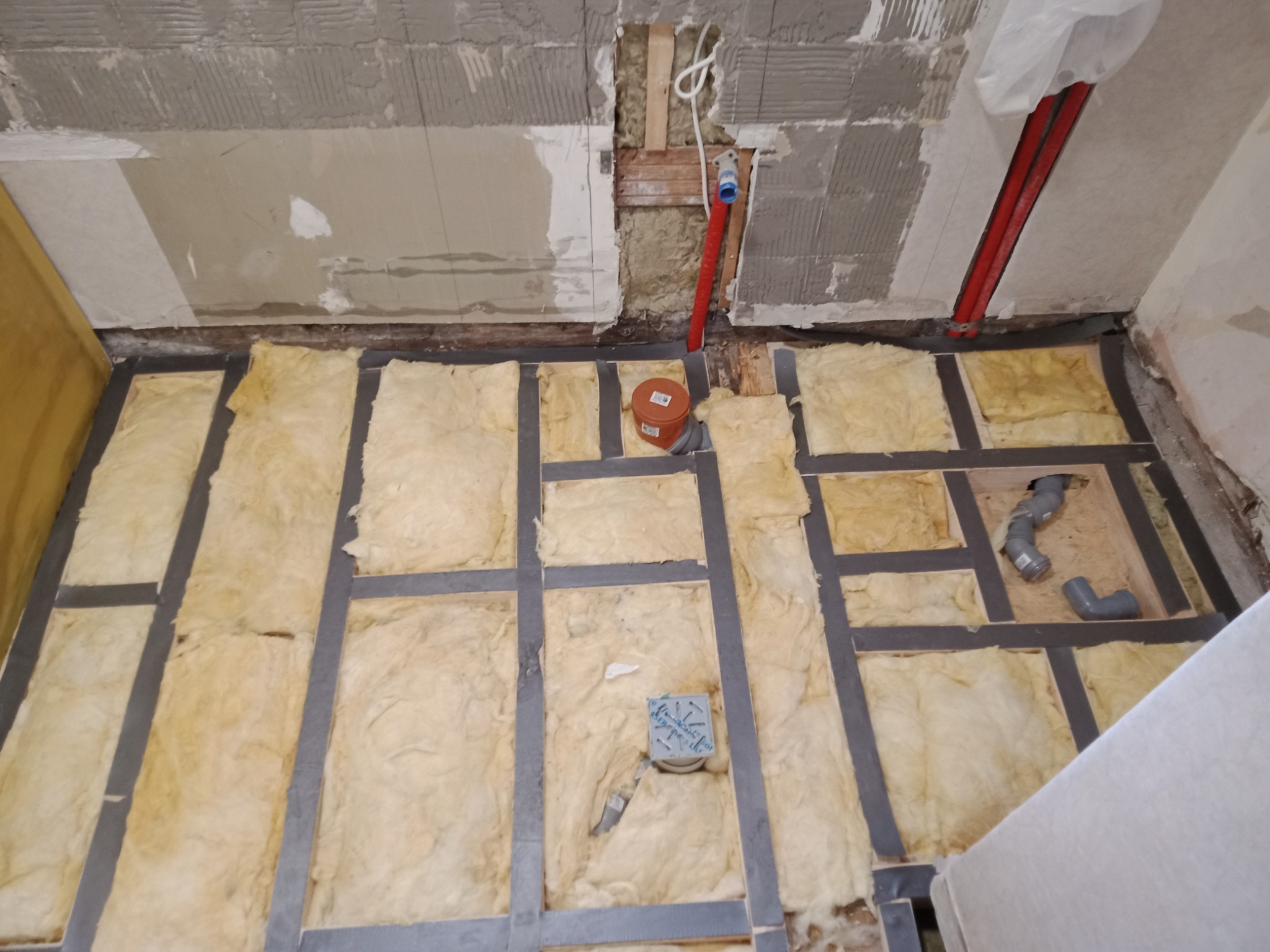 Przygotowanie podłoża pod podłogę: izolacja z wełny mineralnej między legarami, widoczne rury instalacji wodnej i kanalizacyjnej, surowy stan ścian.