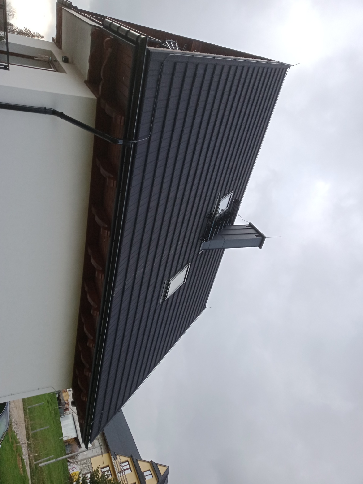 Ciemnoszary dach z blachy na nowoczesnym domu z oknami dachowymi i orynnowaniem, widziany z dołu pod pochmurnym niebem.