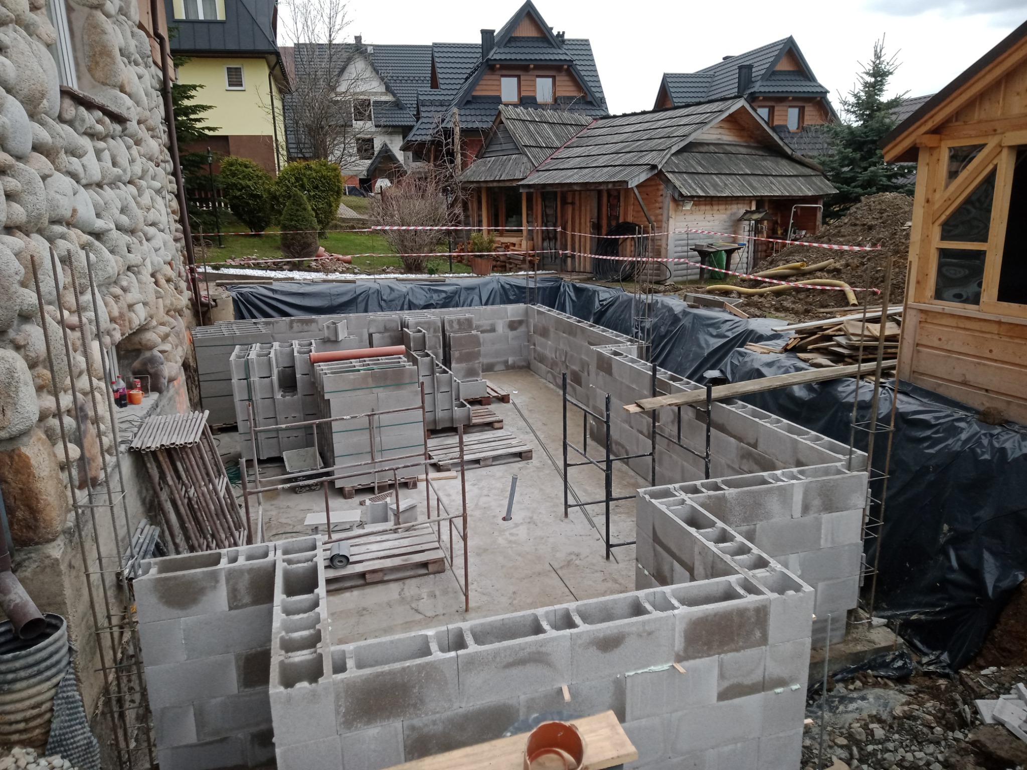 Budowa fundamentów z bloczków betonowych na posesji z zabudową w stylu zakopiańskim, widoczne zbrojenia i elementy szalunków.