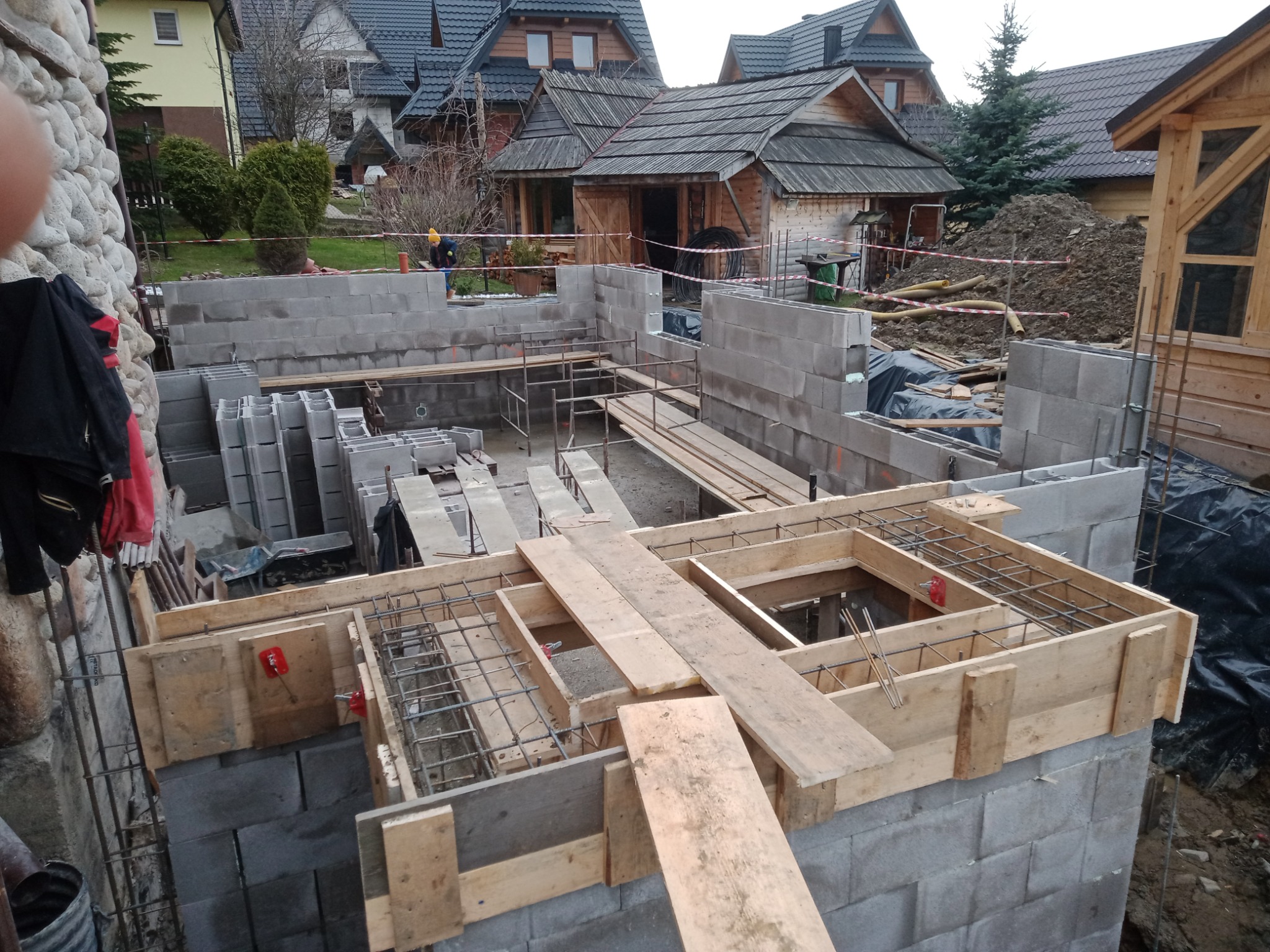 Budowa fundamentów domu jednorodzinnego z bloczków betonowych, widoczne szalunki i zbrojenie, w tle zabudowa w stylu góralskim.