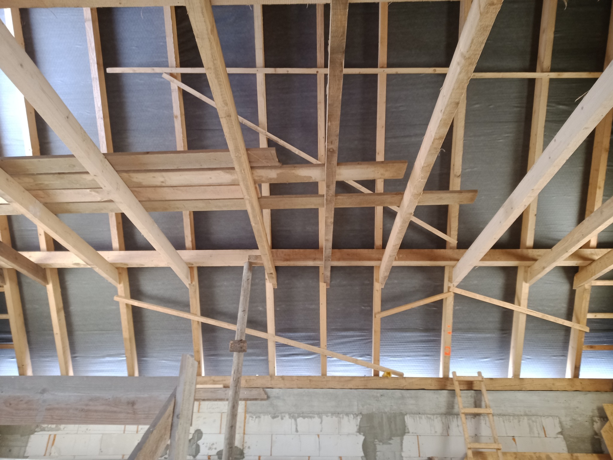 Konstrukcja drewniana dachu w trakcie budowy, widok od wewnątrz, z widocznymi elementami więźby i deskowaniem pokrytym membraną, ściany z betonu komórkowego w dolnej części kadru.