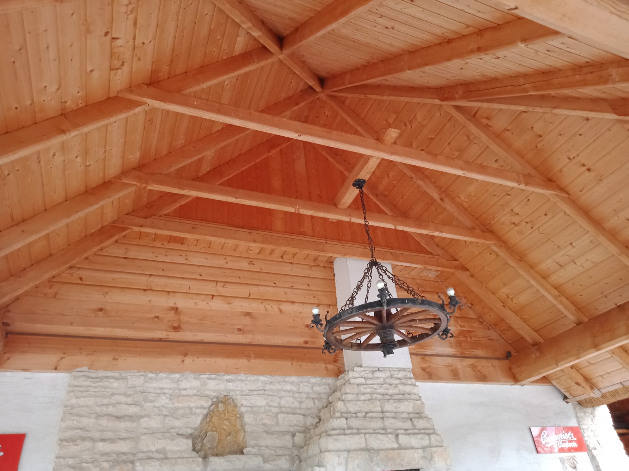 Drewniany sufit z widocznymi belkami i dekoracyjnym żyrandolem w kształcie koła od wozu, ściana z kamienia z dekoracyjnym wnęką.