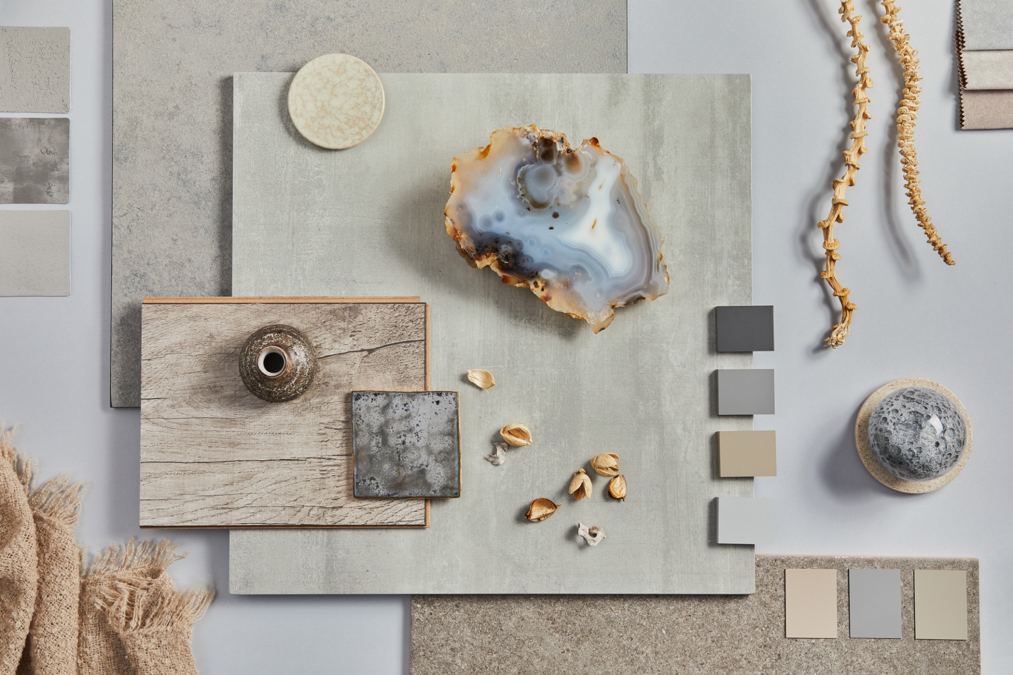 Prezentacja próbek materiałów wykończeniowych: płytki ceramiczne w odcieniach szarości, drewno, kamień agatowy, dekoracyjna gałązka, próbki tkanin i kolorów farb, tworzących paletę barw i tekstur...