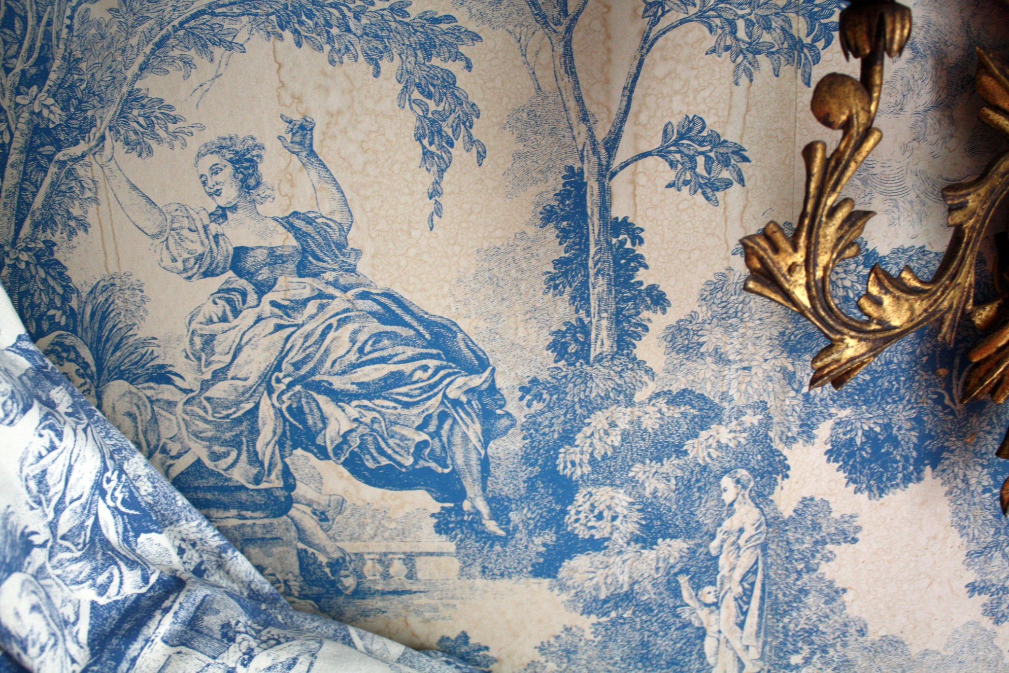 Fragment tapety z motywem toile de Jouy w odcieniach błękitu, przedstawiająca sceny parkowe z postaciami, uzupełniona fragmentem złotej, dekoracyjnej ramy kinkietu.