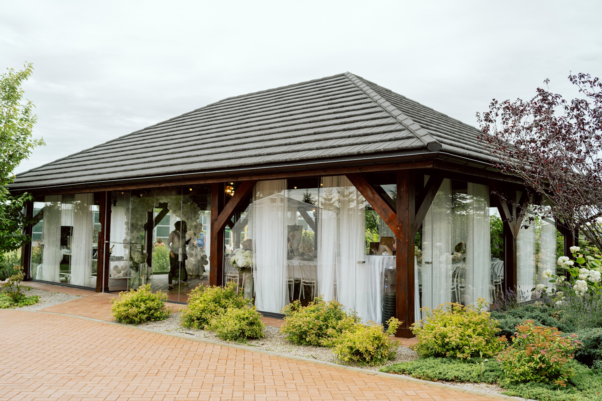 Przeszklona altana z ciemnobrązową drewnianą konstrukcją i szarym dachem z dachówki, udekorowana białymi zasłonami, balonami i kwiatami na przyjęcie, widoczna z zewnątrz na tle ceglanej ścieżki...