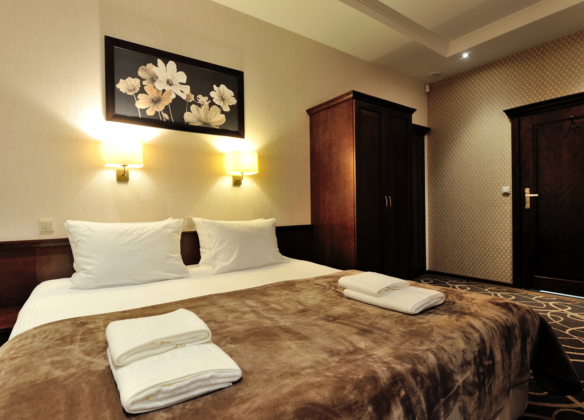 Pokój hotelowy z brązowym pledem na łóżku, białymi ręcznikami i obrazem kwiatów na ścianie w ciepłym oświetleniu