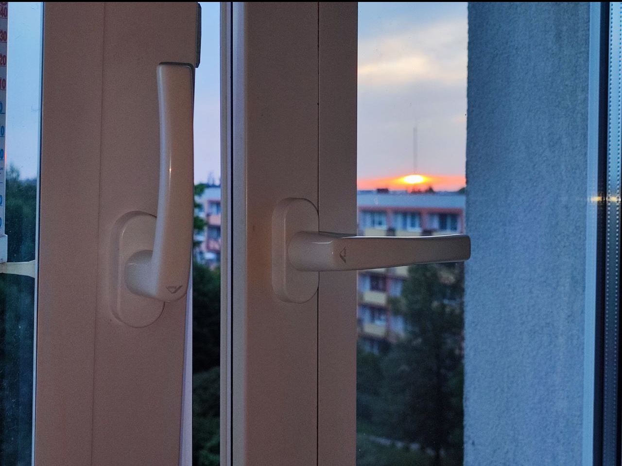 Zamknięte białe okno PCV z widokiem na budynek i zachodzące słońce, widoczny termometr przy ramie okiennej.