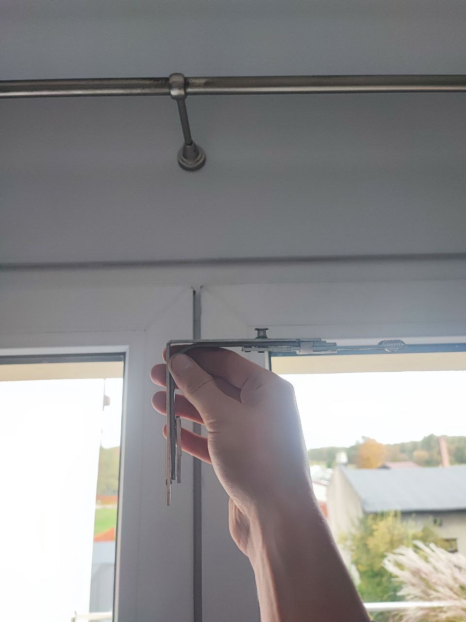 Ręka trzyma metalowy uchwyt karnisza okiennego na tle okna z widokiem na budynki.