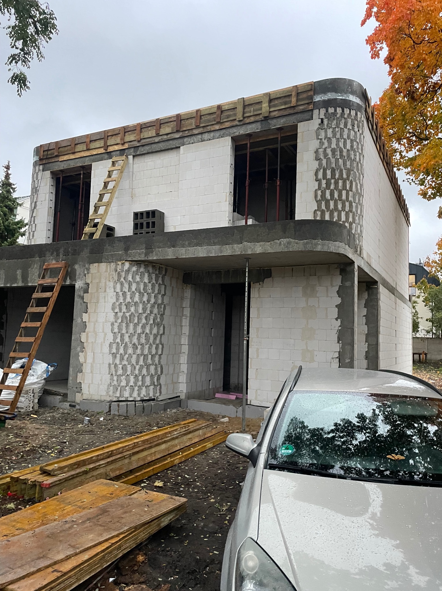 Budowa domu z białych bloczków, widoczne rusztowania i drewniane szalunki. Charakterystyczny wzór na elewacji. Częściowo widoczny samochód na pierwszym planie.